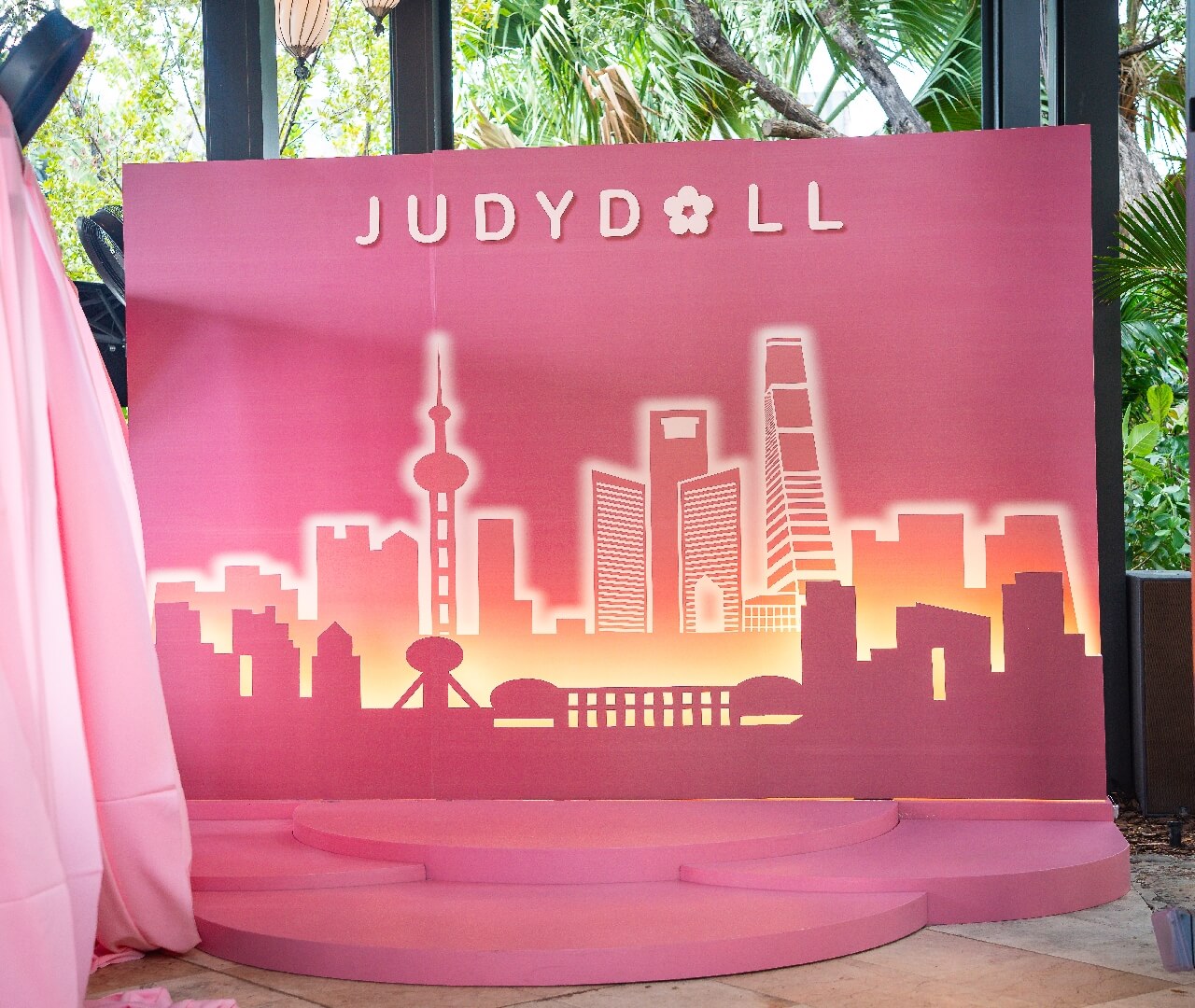 Custom Brand Activation Stage Silhouette Backdrop Custom-designed event production stage featuring a layered pink city skyline silhouette backdrop with warm integrated lighting, dimensional brand lettering, and a tiered circular floor platform built for high-end corporate functions and event rentals.